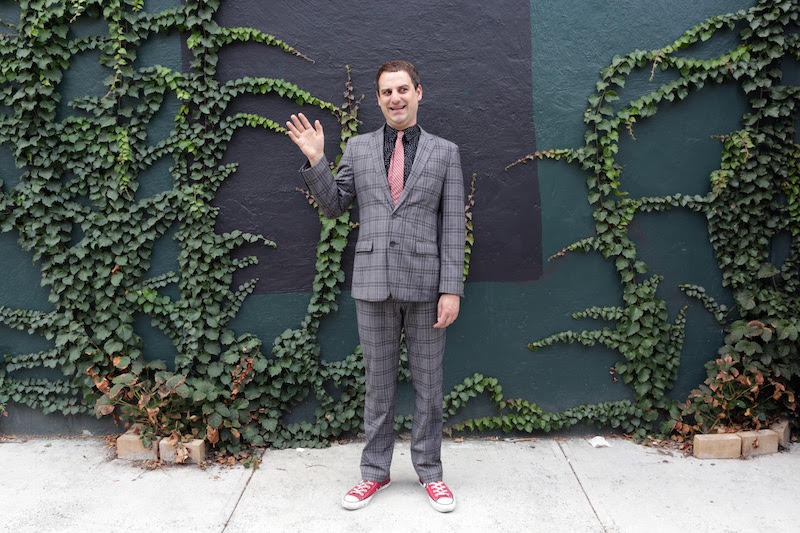 Jeff Seal in a grey plaid suit waves awkwardly to the camera